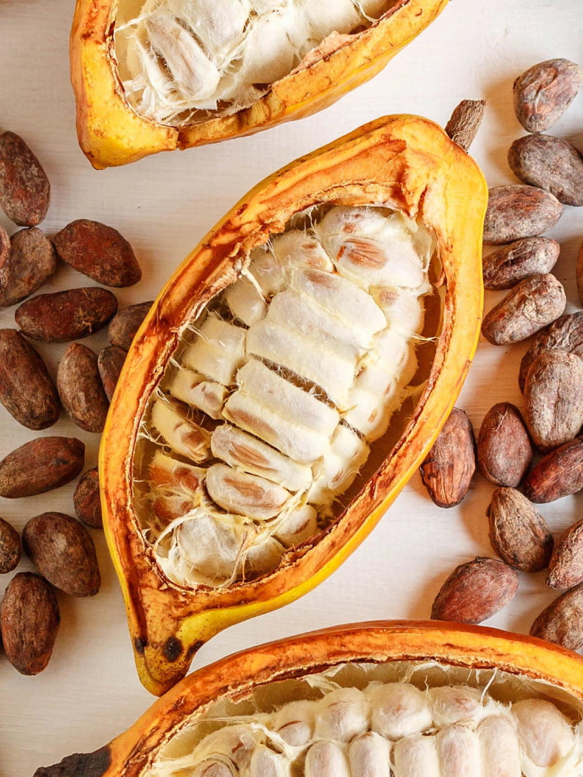 Halved yellow cacao pods with white pulp, dried cacao beans and green leaves on a pale wooden surface.
