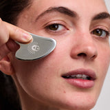 Viva Maia stainless steel Gua Sha tool gliding under the eye of a woman with dark hair tied back.